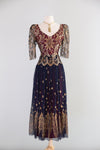 A vintage two-tone dress by Zandra Rhodes displayed on a mannequin against a light gray background. The bodice is burgundy with gold floral embroidery and sheer patterned sleeves, while the skirt is navy blue with gold floral motifs and a gold embroidered border.