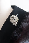 A silver-toned floral brooch set with clear crystals, displayed on a black velvet surface next to a dark fur trim.