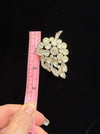 A vintage rhinestone brooch shaped like a grapevine cluster, shown next to a pink measuring tape held by a hand against a black background.