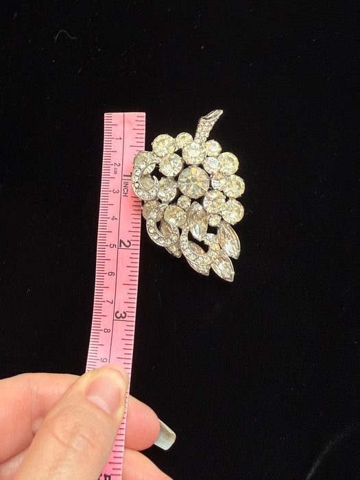 A vintage rhinestone brooch shaped like a grapevine cluster, shown next to a pink measuring tape held by a hand against a black background.